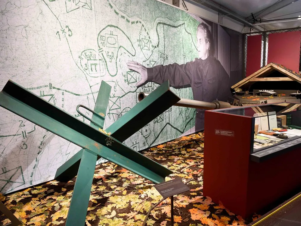 Ausstellungsraum: Karte an der Wand, grüne Metallstrukturen und Informationsstand auf einem Blätterboden