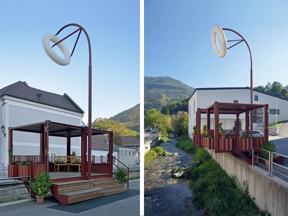 Zwei Ansichten einer roten Struktur mit weißem Ring und überdachtem Bereich, links mit Sitzbank und Pflanzen vor einem Gebäude, rechts über einem Fluss mit Bergblick.