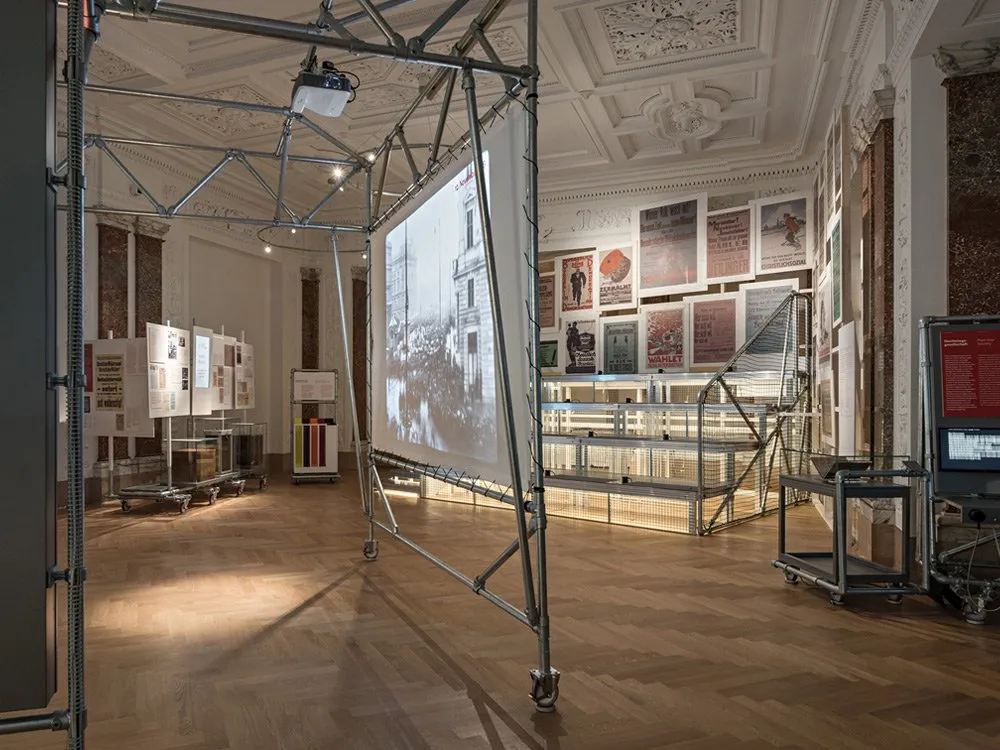 Ausstellungsraum: Metallkonstruktion mit Leinwand zeigt ein Schwarz-Weiß-Bild, umgeben von Vitrinen und Plakaten an den Wänden eines historischen Raums mit Holzboden