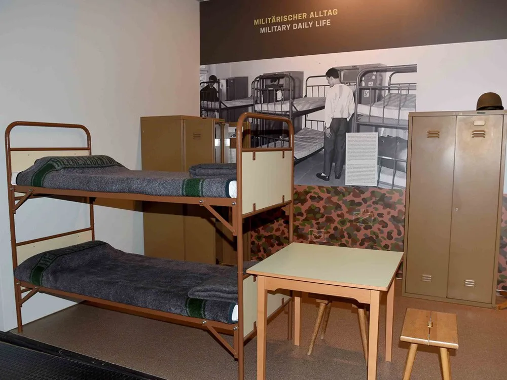 Ausstellungsraum: Etagenbetten, Tisch, Schrank und Foto von militärischem Alltag an der Wand