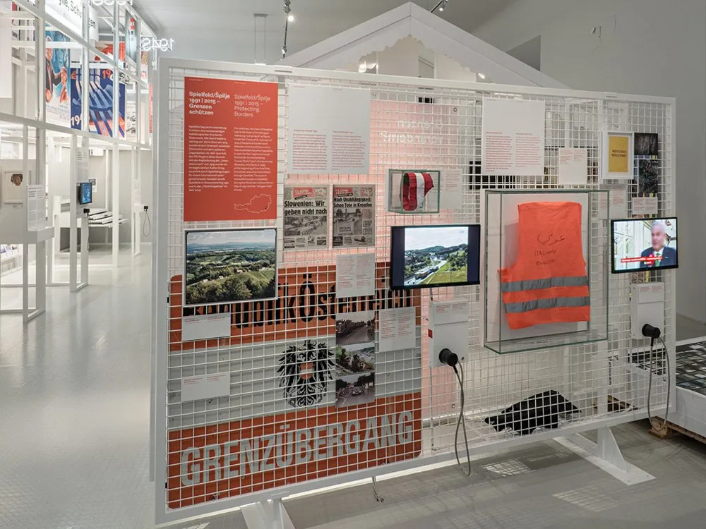 Ausstellungsraum: Wand mit Schild "Grenzübergang" zeigt Fotos, Dokumente und eine orangefarbene Warnweste, umgeben von Vitrinen und Bildschirmen mit historischen Aufnahmen