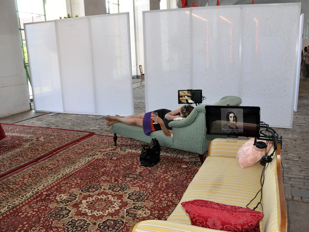 Ausstellungsraum mit einer Person auf einem antiken Sofa mit Kopfhörern und Bildschirm, umgeben von Teppichen und Trennwänden