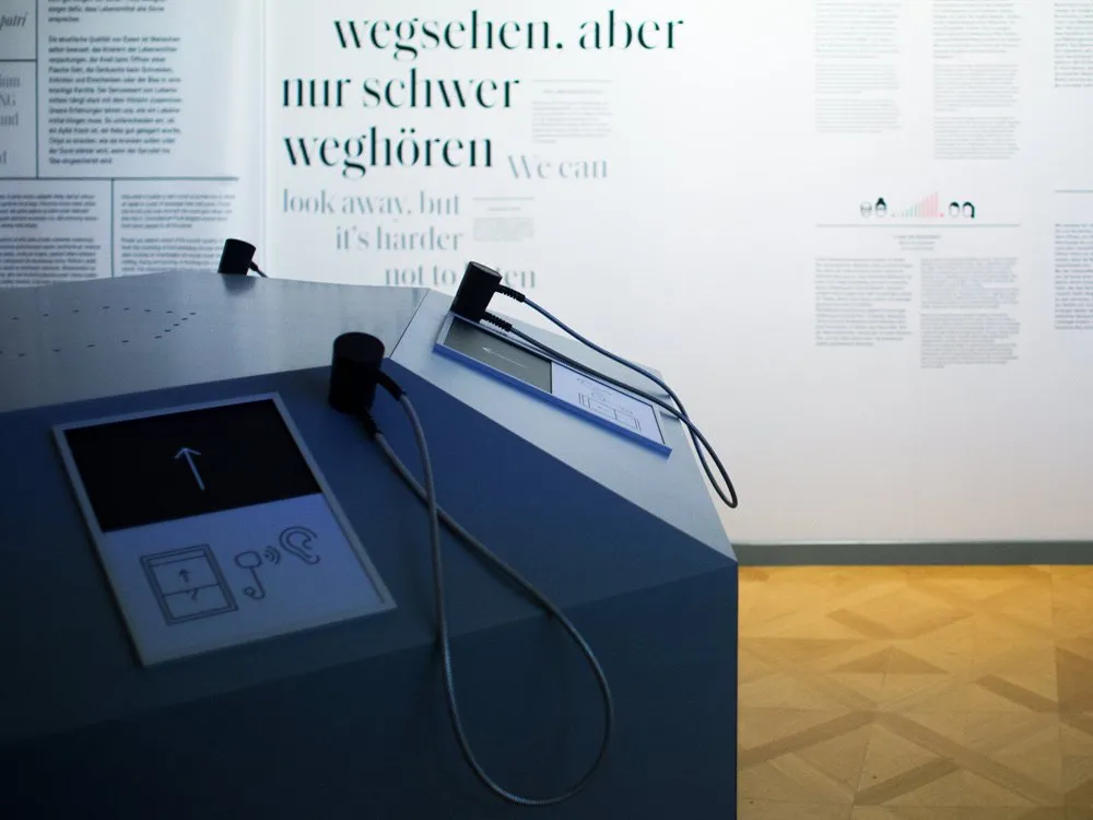 interaktiver Bildschirm mit Kopfhörern, vor einer Wand mit dem Text "Wegsehen, aber nur schwer weg hören"