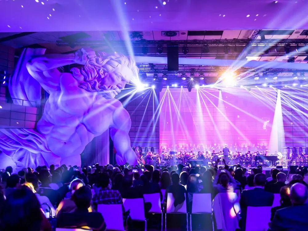 Saal mit übergroßen Poseidon Figur, Orchester auf der Bühne und Publikum auf weißen Stühlen, beleuchtet von bunten Lichtern.