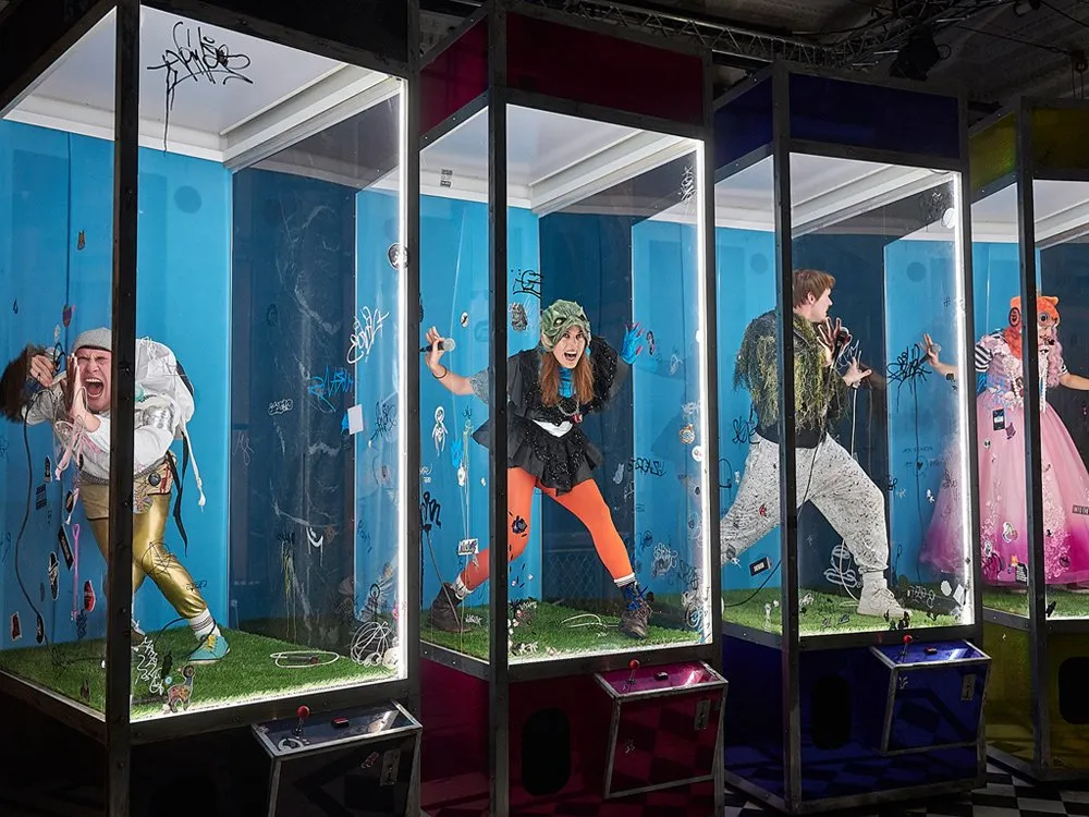 Vier bunte Glasvitrinen mit grasbedecktem Boden, jede mit einer Person in auffälliger Kleidung, die Mikrofone hält, umgeben von Graffiti und Stickern, mit Joysticks an der Basis