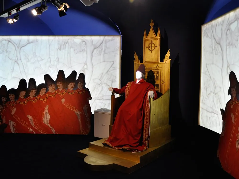 Ausstellungsraum: Figur in roten Gewändern sitzt auf goldenem Thron, umgeben von projizierten Bildern roter Figuren in Kapuzenmänteln an den Wänden