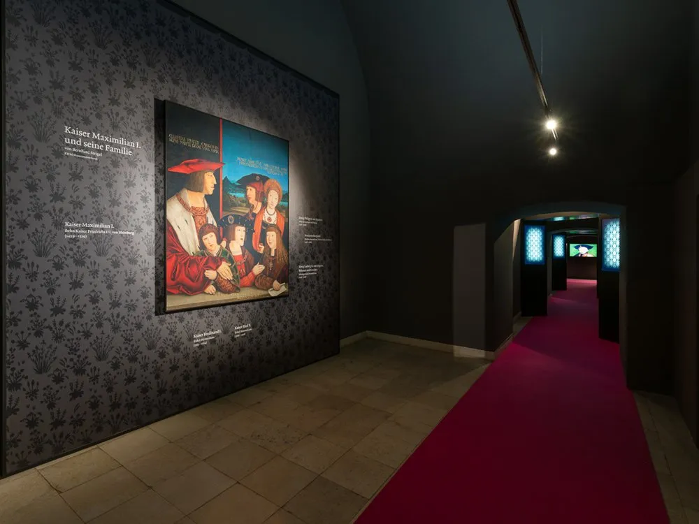 Ausstellungsraum: Düsterer Raum mit rotem Teppich, Gemälde von Kaiser Maximilian I. und seiner Familie sowie beleuchtete Vitrinen
