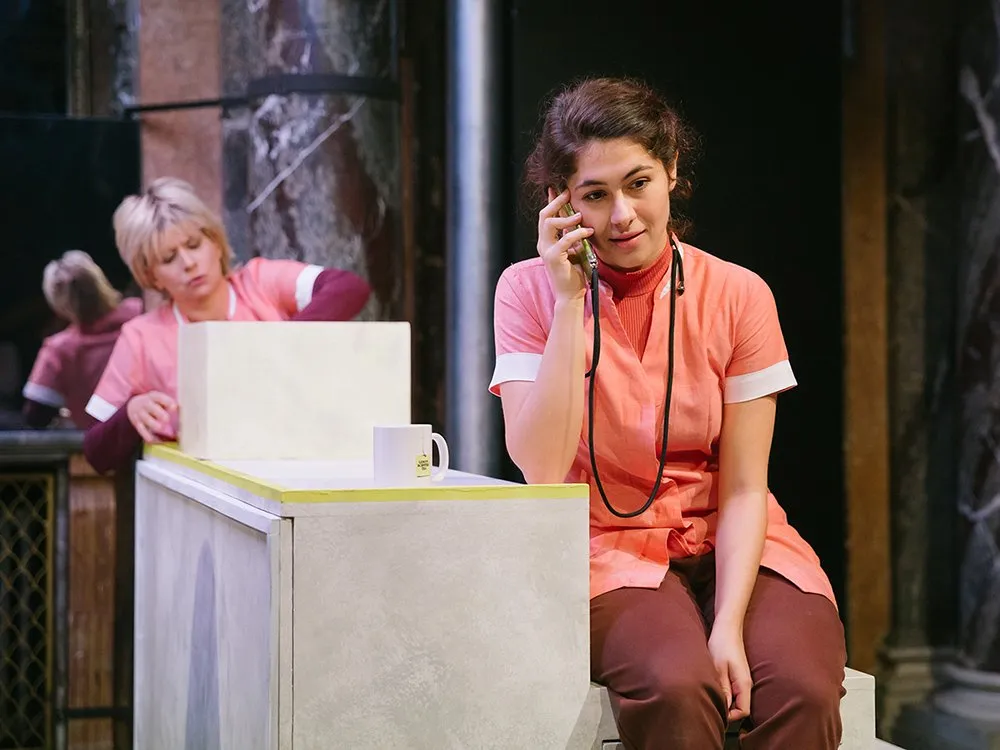 Schauspielerin in orange-uniformartigem Outfit telefoniert an einem weißen Tisch mit Tasse, im Hintergrund eine weitere Person an einem Tisch, umgeben von marmorierten Säulen