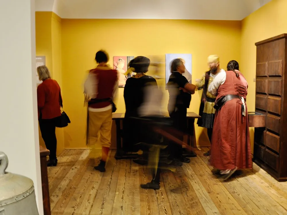 Gelber Ausstellungsraum mit Holzboden, wo mehrere Menschen Gemälde an der Wand betrachten