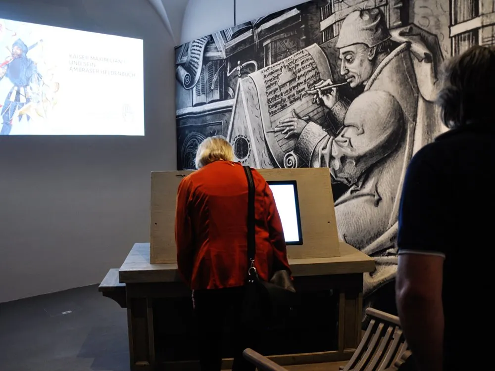 Person steht vor Bildschirm im Ausstellungsraum, historische Zeichnung eines schreibenden Mannes an der Wand und ein Projektor mit „Kaiser Maximilian I. und sein Ambraser Heldenbuch“