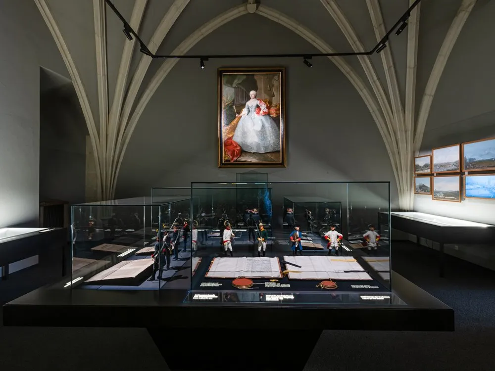 Ausstellungsraum: Gotischer Raum mit hohem Gewölbe, einem Porträt an der Wand und einer Vitrine mit Miniaturfiguren und historischen Dokumenten