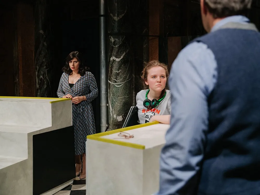 Junge Schauspielerin, mit Kopfhörern um den Hals, sitzt an einem weißen Tisch, mit ernstem Blick an Mann im Vordergrund