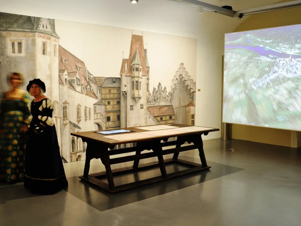 Ausstellungsraum mit einer historischen Wandmalerei von mittelalterlichen Gebäuden, einem Holztisch mit Dokumenten und zwei Personen in historischen Kostümen