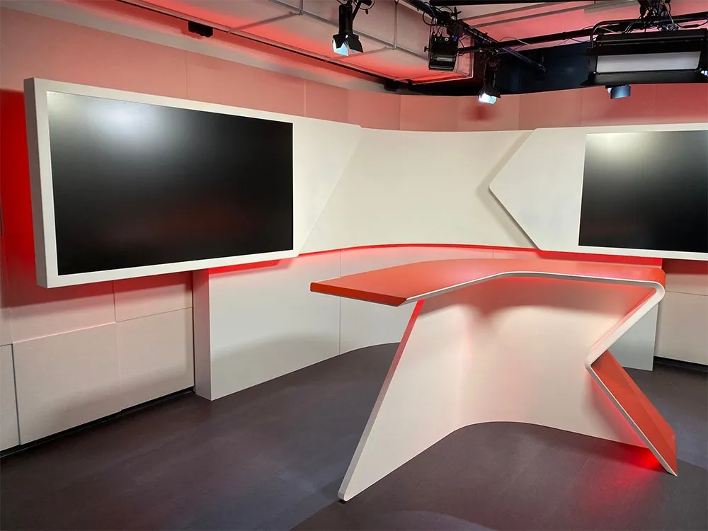 Nachrichtenstudio-Set mit rot-weißen Elementen, zwei großen schwarzen Bildschirmen und einem futuristischen Pult