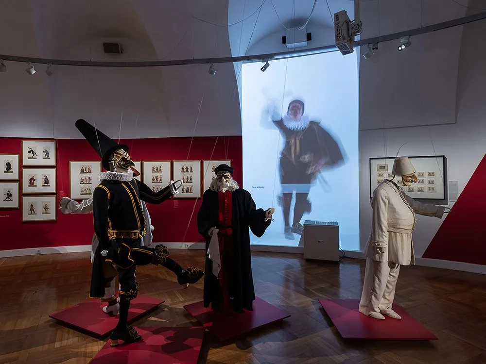 Ausstellungsraum: Drei Marionetten in historischen Kostümen auf roten Podesten, eine mit schwarzem Hut und goldener Maske. Hintergrund mit Projektion und Zeichnungen an roter Wand.