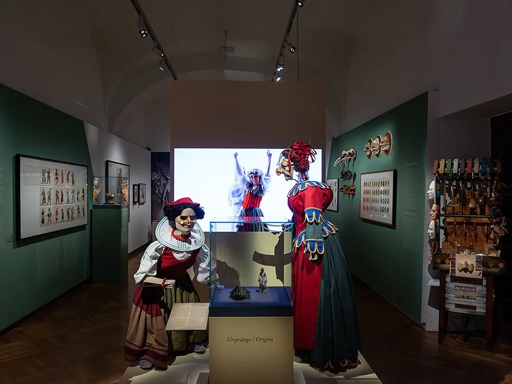 Ausstellungsraum: Drei Marionetten in bunten historischen Kostümen, eine mit roter Kleidung und roter Haube, vor einer Projektion. Im Hintergrund gerahmte Zeichnungen und Vitrinen an grünen Wänden.