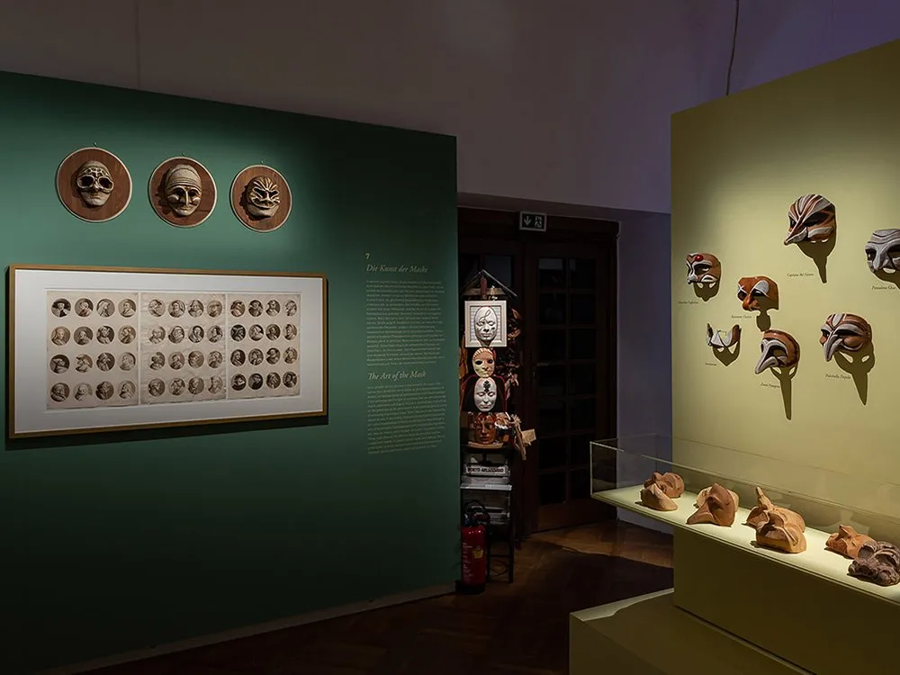 Ausstellungsraum: Drei Masken an einer grünen Wand, ein gerahmtes Bild mit vielen Porträts darunter. Rechts gelbe Wand mit weiteren Masken