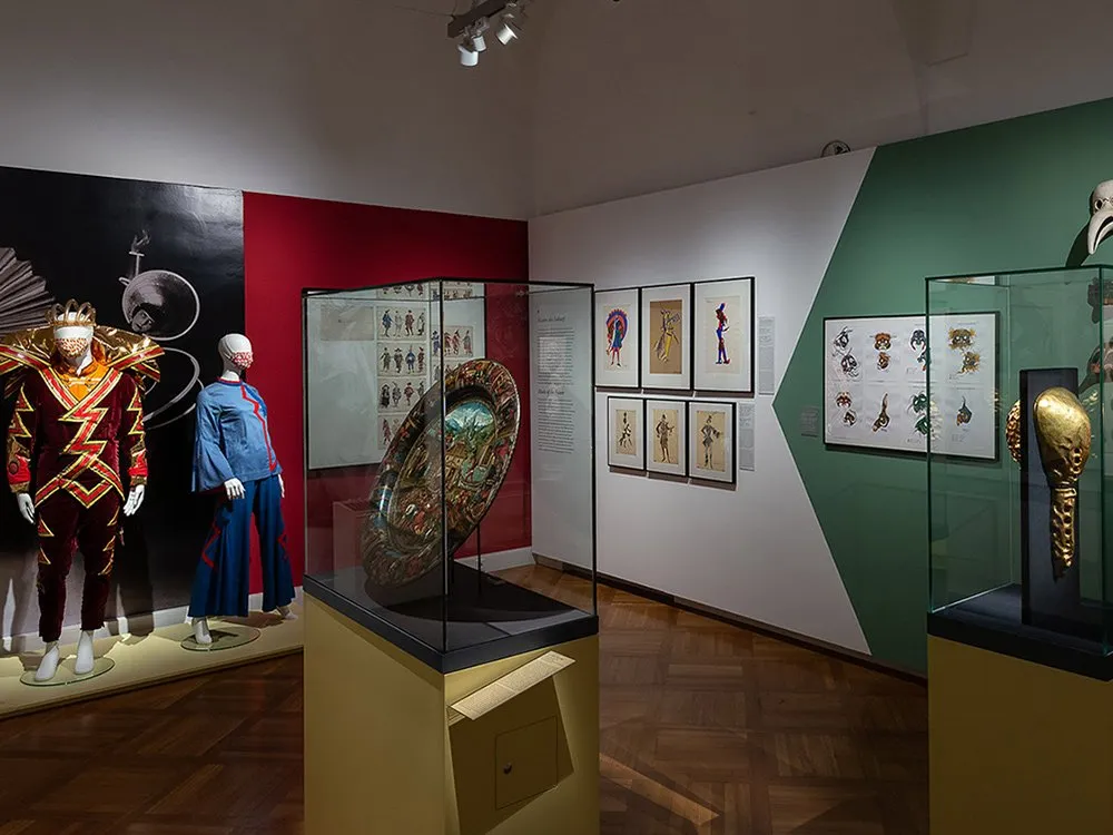 Ausstellungsraum: Zwei Schaufensterpuppen mit farbigen Kostümen, eine Vitrine mit einem bemalten Schild, gerahmte Zeichnungen an den Wänden und goldene Maske in einer Vitrine.