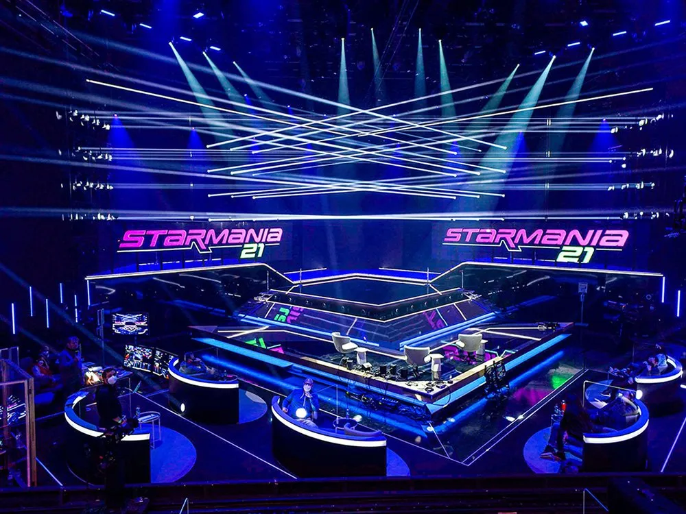 Moderne Bühne in blauem Licht, durchzogen von weißen Laserstrahlen, die sich kreuzen. Die mehrstufige Fläche mit leuchtenden Rändern trägt das "Starmania 21" Logo in Pink und Grün. Techniker bedienen runde Pultstationen mit Monitoren