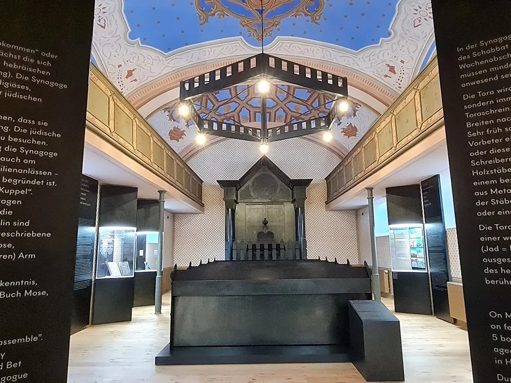 Ausstellungsraum mit einem zentralen schwarzen Altar und bemalter Decke, umgeben von Vitrinen mit Exponaten.