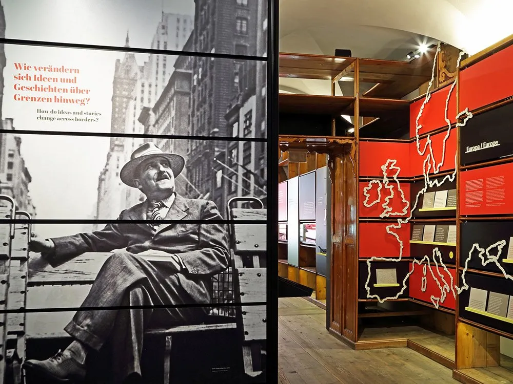 Ausstellungsraum: Schwarz-Weiß-Foto von Stefan Zweig in einem Anzug auf einer Feuerleiter vor Gebäuden, daneben eine rote Wand mit Karten von Europa und Texttafeln über Ideen und Grenzen.
