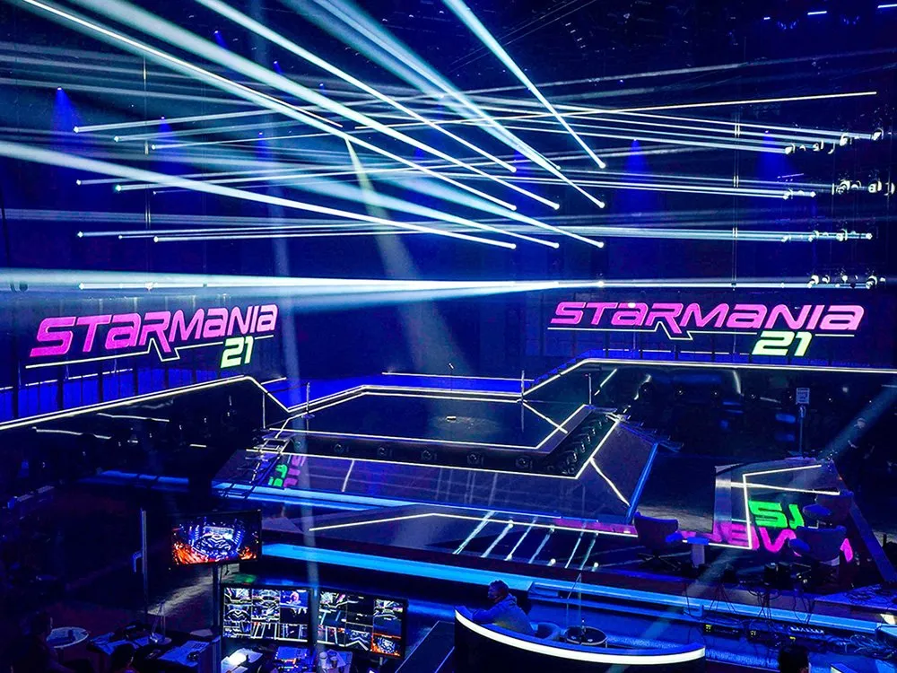 Eine futuristische Bühne mit blauen Lichtern und weißen Laserstrahlen, die sich kreuzen. Die mehrstufige Plattform mit leuchtenden Kanten zeigt das Logo "Starmania 21" in Pink und Grün. Im Vordergrund sind Techniker an Kontrollpulten mit Monitoren zu sehen