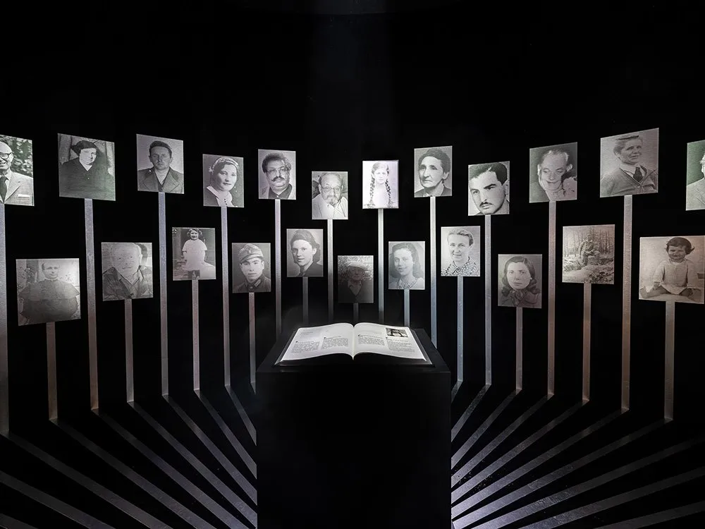 Dunkler Ausstellungsraum mit einem offenen Buch auf einem Podest, umgeben von Ständern mit historischen Schwarz-Weiß-Fotografien von Personen.