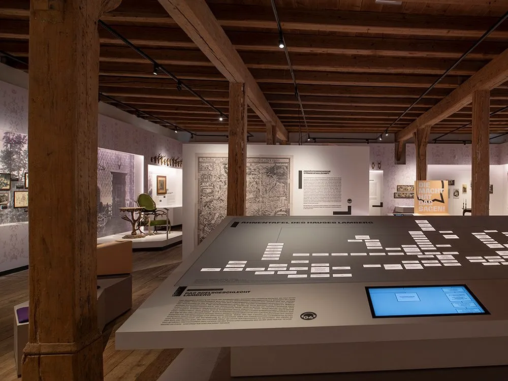 Ausstellungsraum: Im Vordergrund eine interaktive Tafel mit Stammbaum der Familie Lamberg, im Hintergrund historische Gemälde, Fotos und ein grüner Stuhl als Exponat.