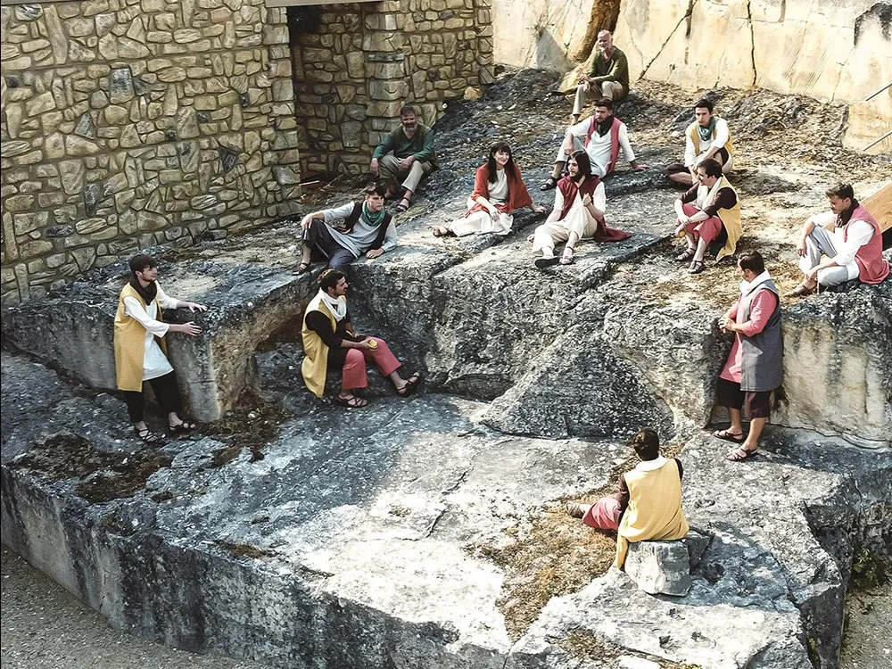 Szene: Darsteller in historischen Gewändern sitzen auf Felsen und unterhalten sich, andere stehen oder gestikulieren, umgeben von einer steinernen Kulisse