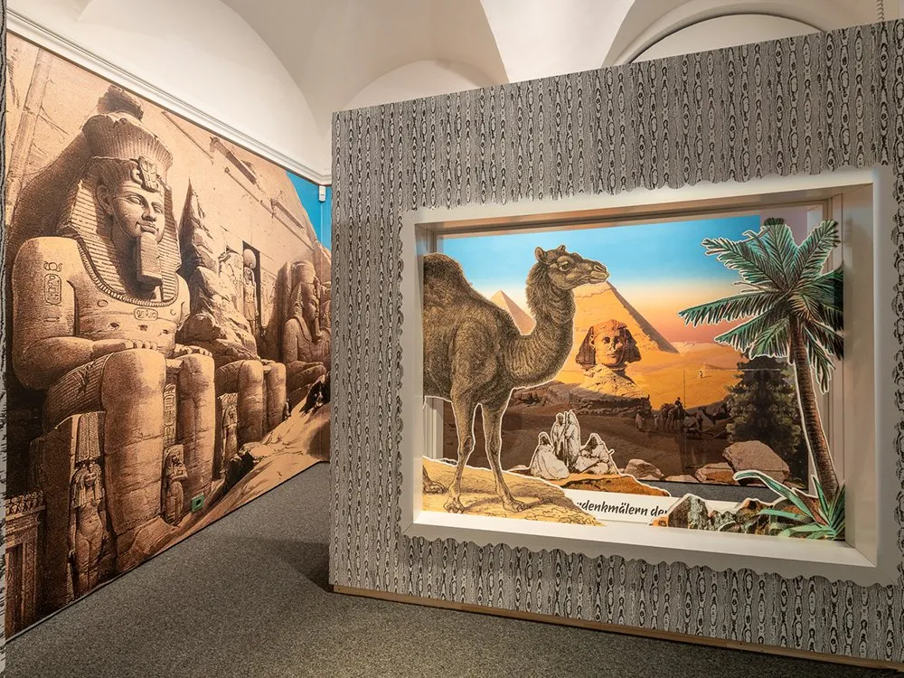 Ausstellungsraum: Wände mit Wandbildern von ägyptischen Statuen und einer Szene mit Kamel, Pyramide und Palmen. Eine Vitrine zeigt historische Artefakte, ergänzt durch eine weitere Darstellung exotischer Landschaften.