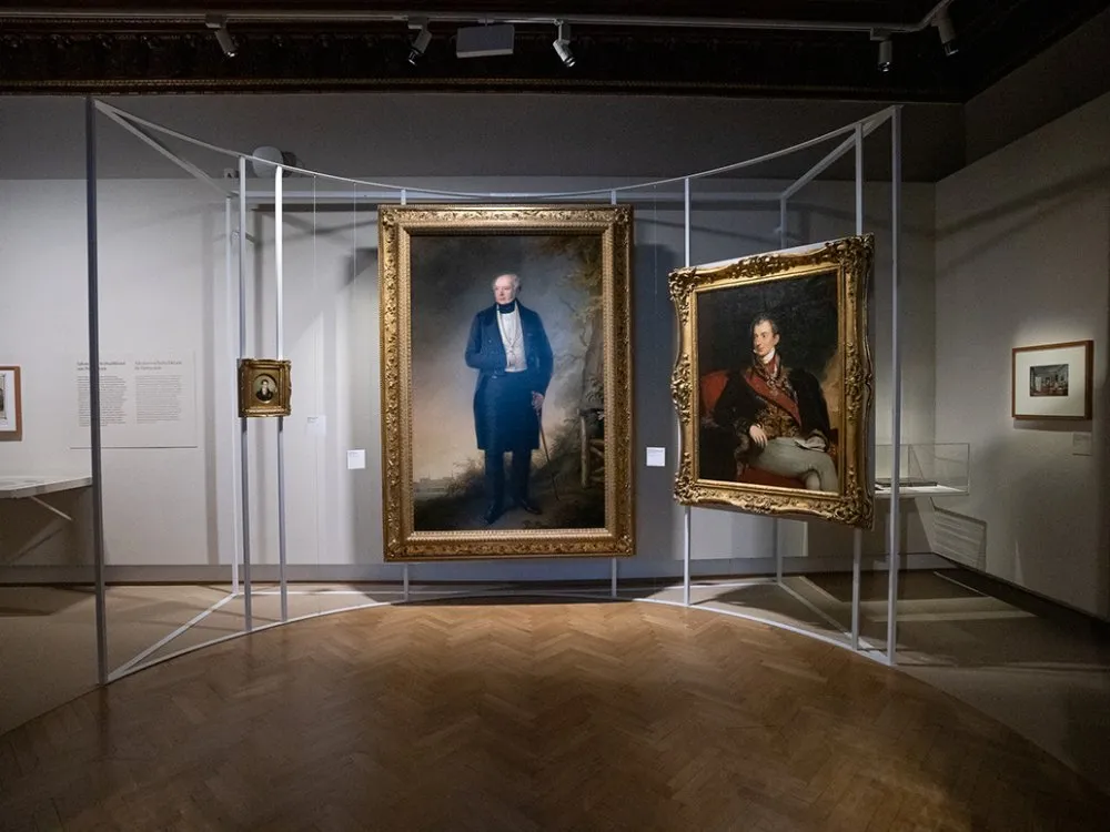 Ausstellungsraum: Zwei große Porträts in goldenen Rahmen von historischen Figuren, umgeben von kleineren Bildern und Texten an der Wand