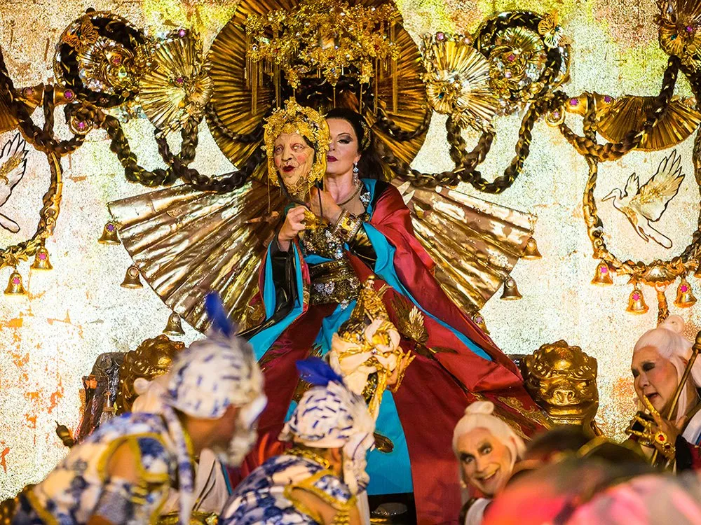 Szene: Eine zentrale Figur in rot-blauem Gewand mit goldener Maske und Krone, umgeben von weiteren Figuren in farbenfrohen Kostümen