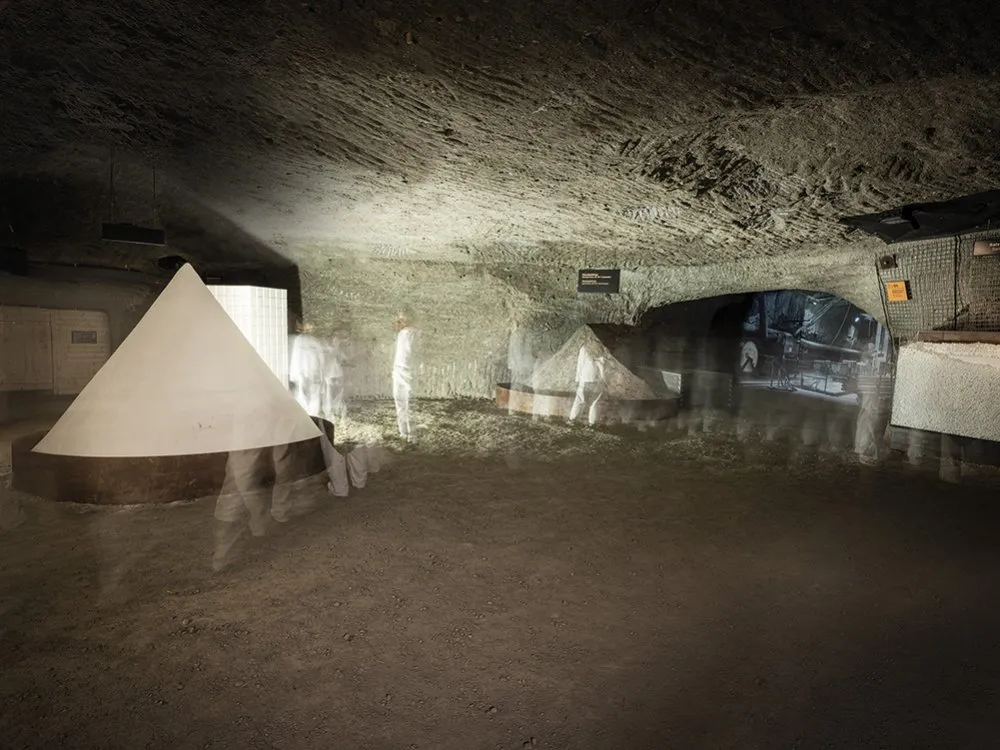 Unterirdischer Ausstellungsraum mit rauer Felsdecke, leuchtenden weißen Pyramidenkegeln auf runden Podesten und leicht transparent sichtbaren Gestalten in weißen Anzügen, aufgenommen mit einer Langzeitbelichtung.
