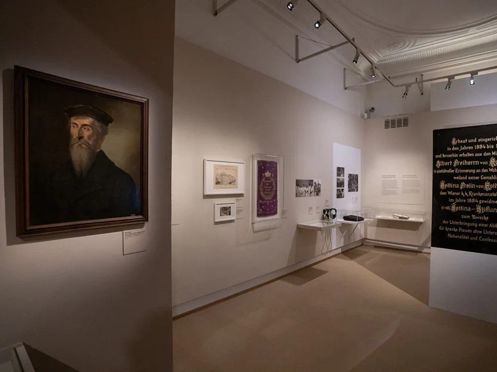 Ausstellungsraum: Historisches Porträt eines bärtigen Mannes in einem dunklen Rahmen, umgeben von alten Dokumenten, Fotos und einem violetten Stoffstück in Vitrinen, mit Texten an der Wand über die Wiener Rothschilds.