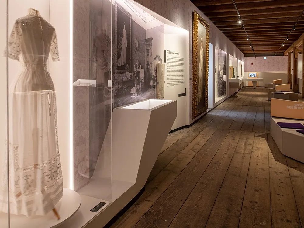 Ausstellungsraum: Vitrinen präsentieren ein historisches Kleid und weitere Artefakte. An den Wänden hängen ein Gemälde einer Frau in Weiß sowie historische Fotos und Infotafeln.