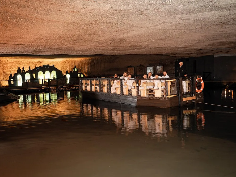 Unterirdischer See mit beleuchtetem Boot, Besucher in weißen Anzügen und einem Führer in dunkler Kleidung, sowie dekorative Fenster an der Felswand