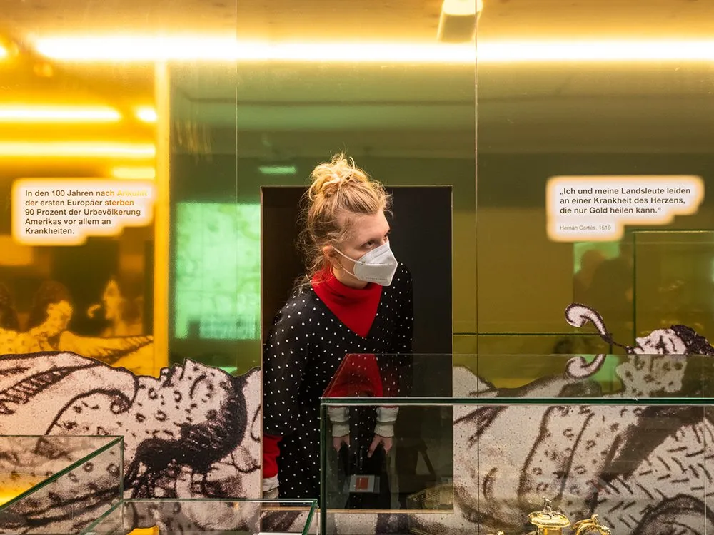 Ausstellungsraum: Eine Person steht vor einer Vitrine mit goldenen Artefakten. Die Wände zeigen Wandbilder mit exotischen Motiven und Texte über historische Ereignisse, darunter Zitate von Hernan Cortés.