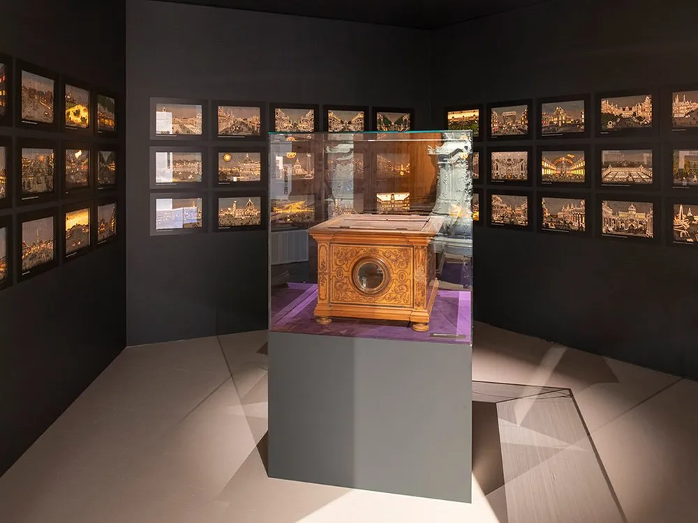 Ausstellungsraum: Zentral platziert eine Vitrine mit einem ornamentierten Holzkiste. An den Wänden hängen zahlreiche Fotos von beleuchteten Stadtansichten.