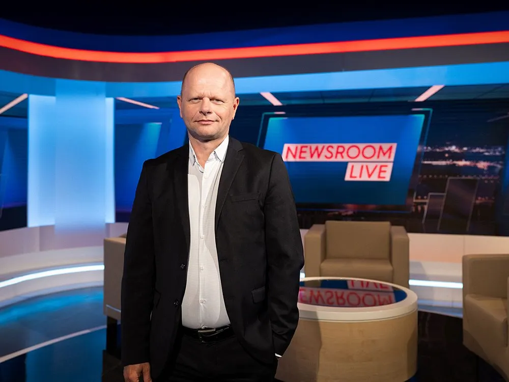 Ein Mann in einem schwarzen Anzug und weißem Hemd steht vor einem modernen Set mit einem runden Tisch und beigen Sesseln, umgeben von blauen und roten Lichtelementen und einem großen Bildschirm mit der Aufschrift "NEWSROOM LIVE".