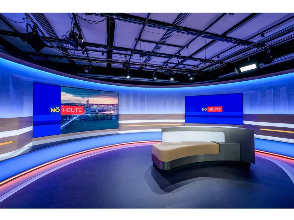 "Niederösterreich heute"-Studio mit gebogener Bühne, LED-Wänden mit Stadtansichten, blauen Akzenten und einem zentralen Pult