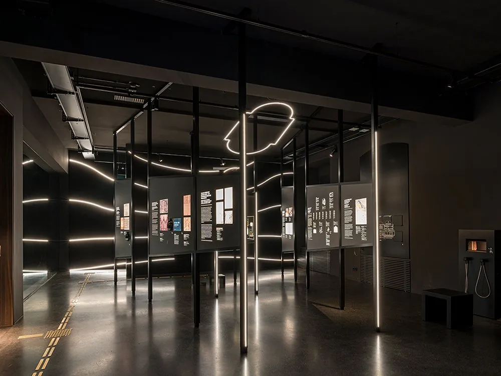 Ausstellungsraum mit schwarzen Panels, beleuchtete Displays mit Bildern und Beschriftungen,  leuchtende Neonlinien an der Wand