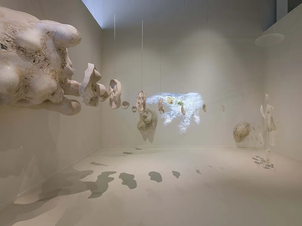 Überlebensgroße Amöbenskulptur und transparente menschliche Fragmente hängen in einem Raum, mit projizierten Mikroorganismen im Hintergrund.