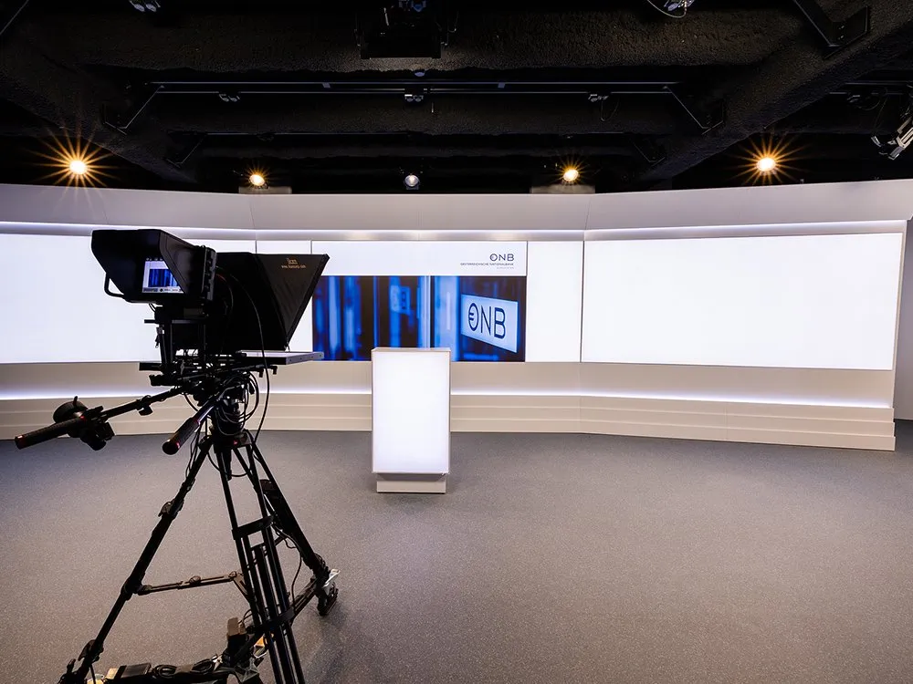 Studio mit einer Kamera auf einem Stativ, einem weißen Pult und einer LED-Wand mit „ÖNB“-Logo