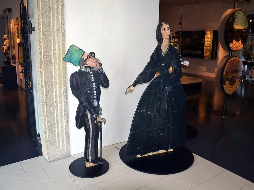 Ausstellungsraum mit zwei historische Figuren, einem Mann in Uniform mit grünem Hut und einer Frau in schwarzem Kleid auf Sockeln, umgeben von Vitrinen und Beschriftungen.