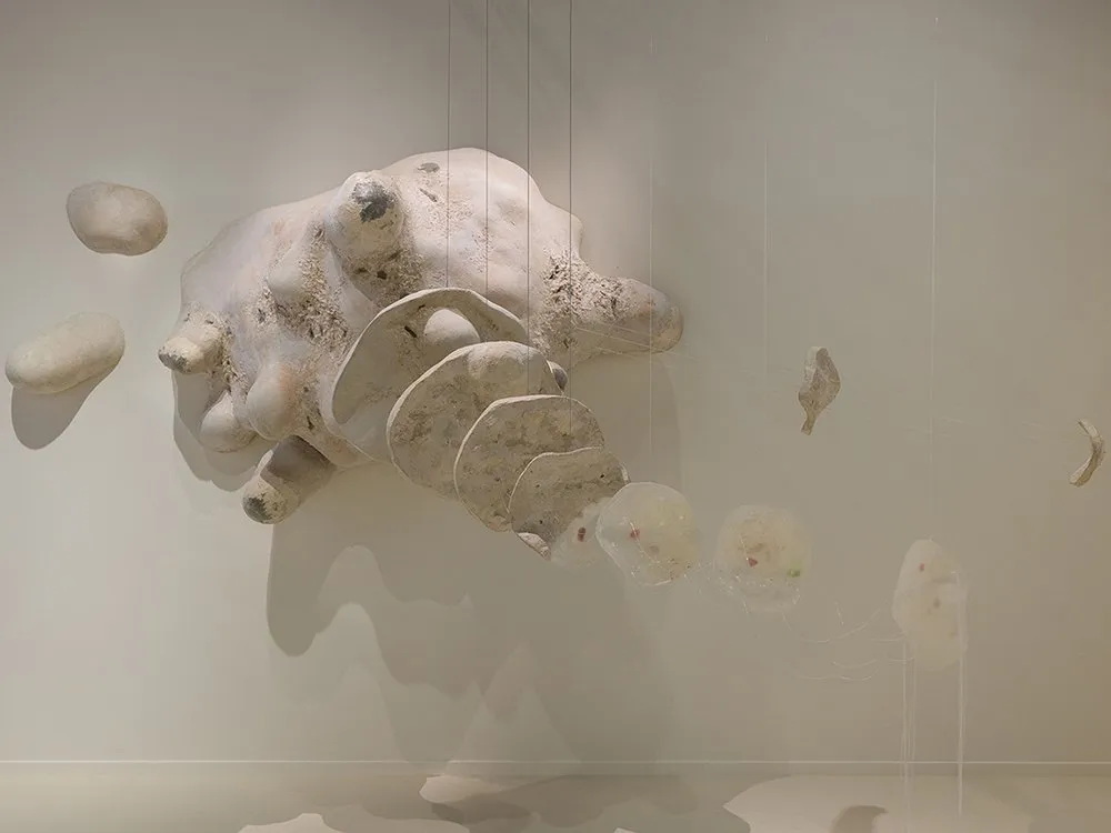 Amöbenskulptur dehnt sich im Raum aus, verknüpft mit durchsichtigen, kugelförmigen Teilen, die Mikroorganismen symbolisieren.