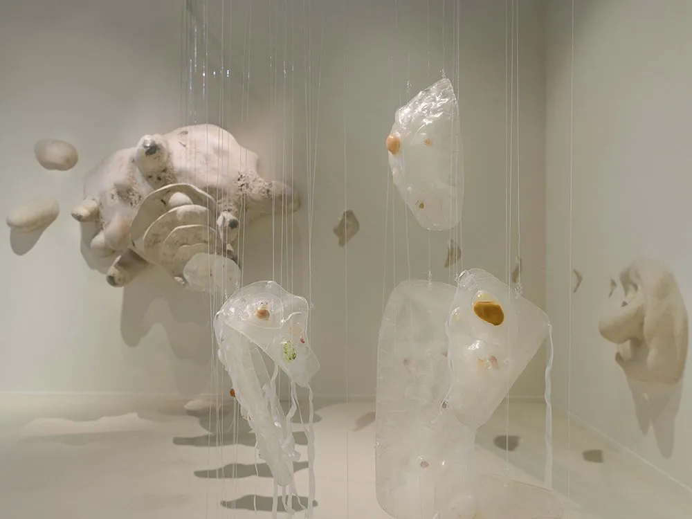 Amöbenskulptur mit ausladenden Formen hängt im Raum, ergänzt durch transparente, kugelförmige Elemente mit eingebetteten Mikroorganismen-Darstellungen.