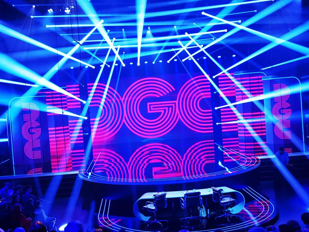 Leuchtende TV-Bühne mit großer LED-Wand im Hintergrund, die den Schriftzug „GGC“ zeigt, davor eine runde Plattform mit einem Tisch und drei Stühlen, durchzogen von Lichtstrahlen.