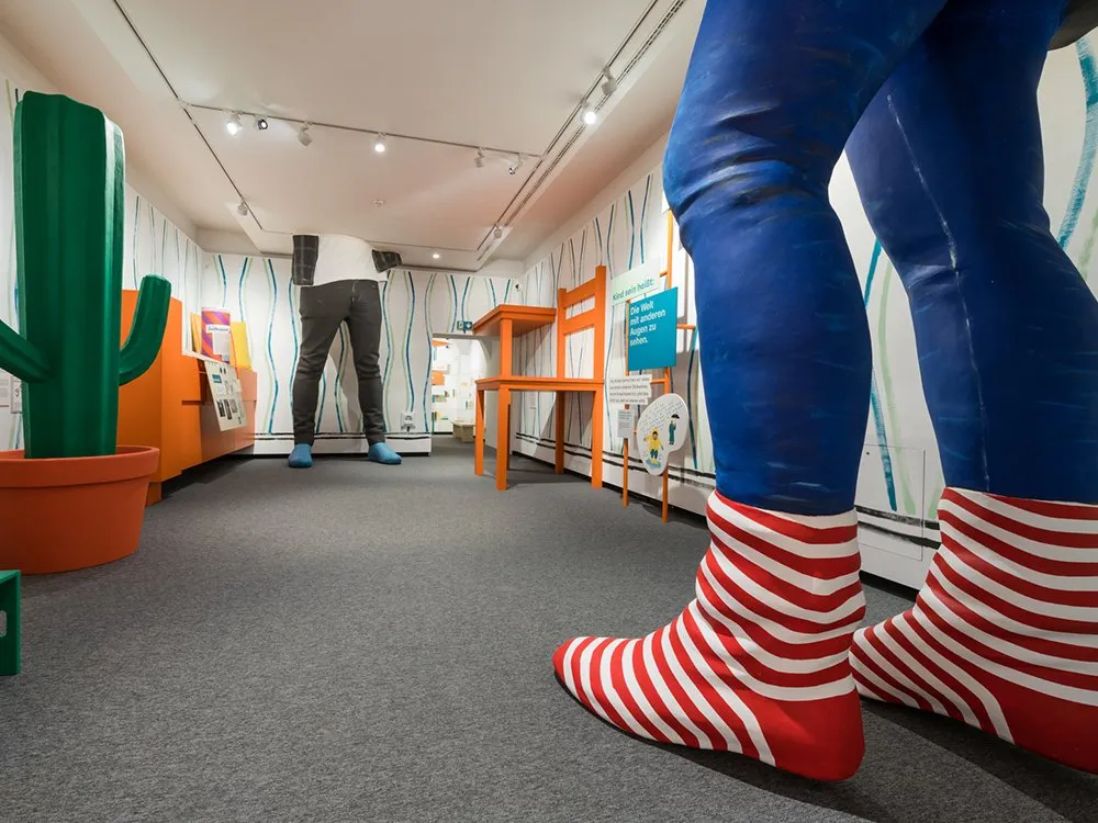 Weitwinkelaufnahme eines Ausstellungsraums mit überlebensgroßen Figuren, eine mit blauen Beinen und roten-weißen Socken, eine mit grauen Hosen und blauen Schuhen, neben einem Kaktus und orangefarbenen Möbeln