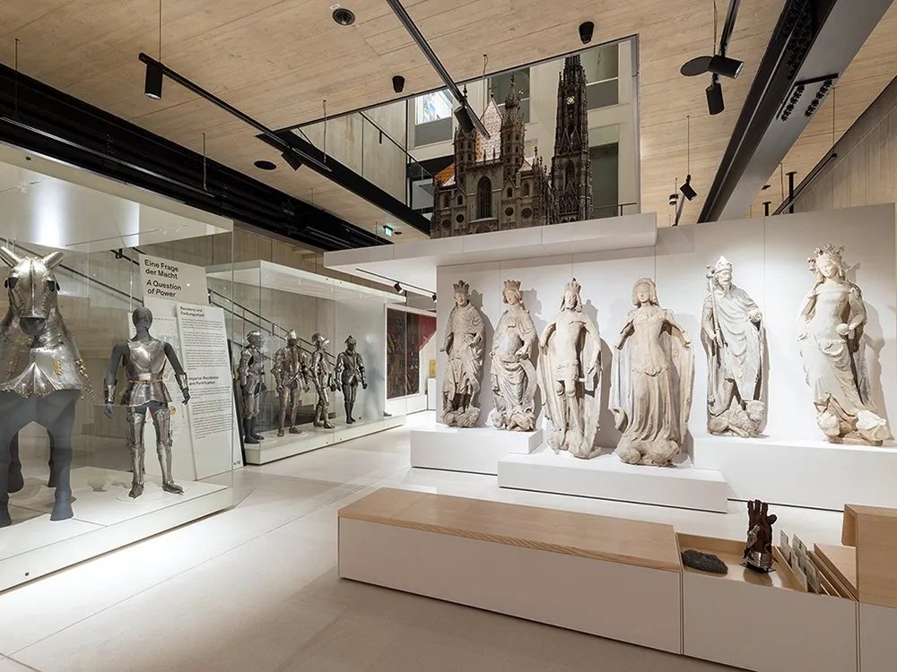 Ausstellungsraum mit Vitrinen, die Ritterrüstungen und historische Statuen zeigen, sowie einem Modell eines gotischen Doms im Hintergrund