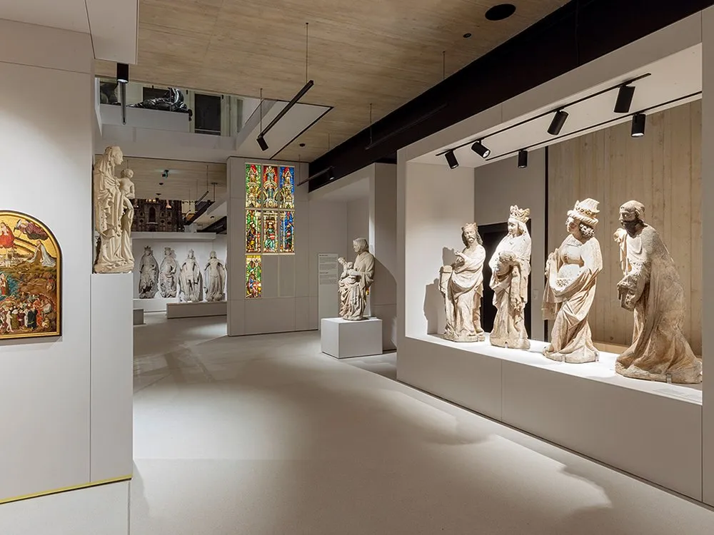 Ausstellungsraum mit Vitrinen, die historische Statuen und ein bunten Kirchenfenstern